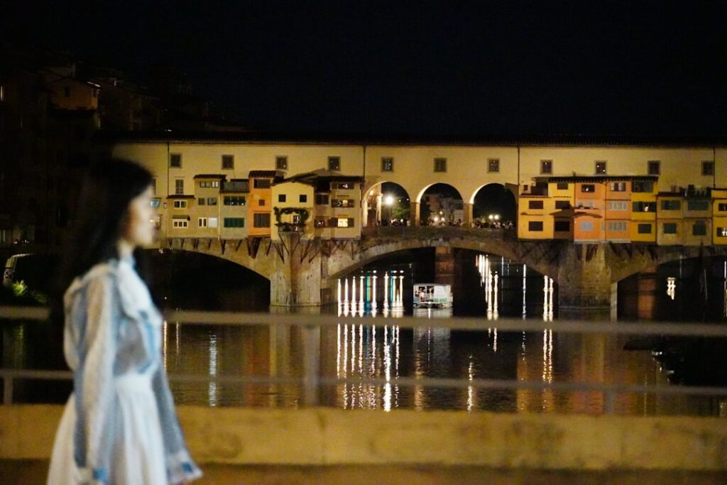 Blurred person walking by the illuminated Ponte Vecchio reflecting on the Arno in Florence.